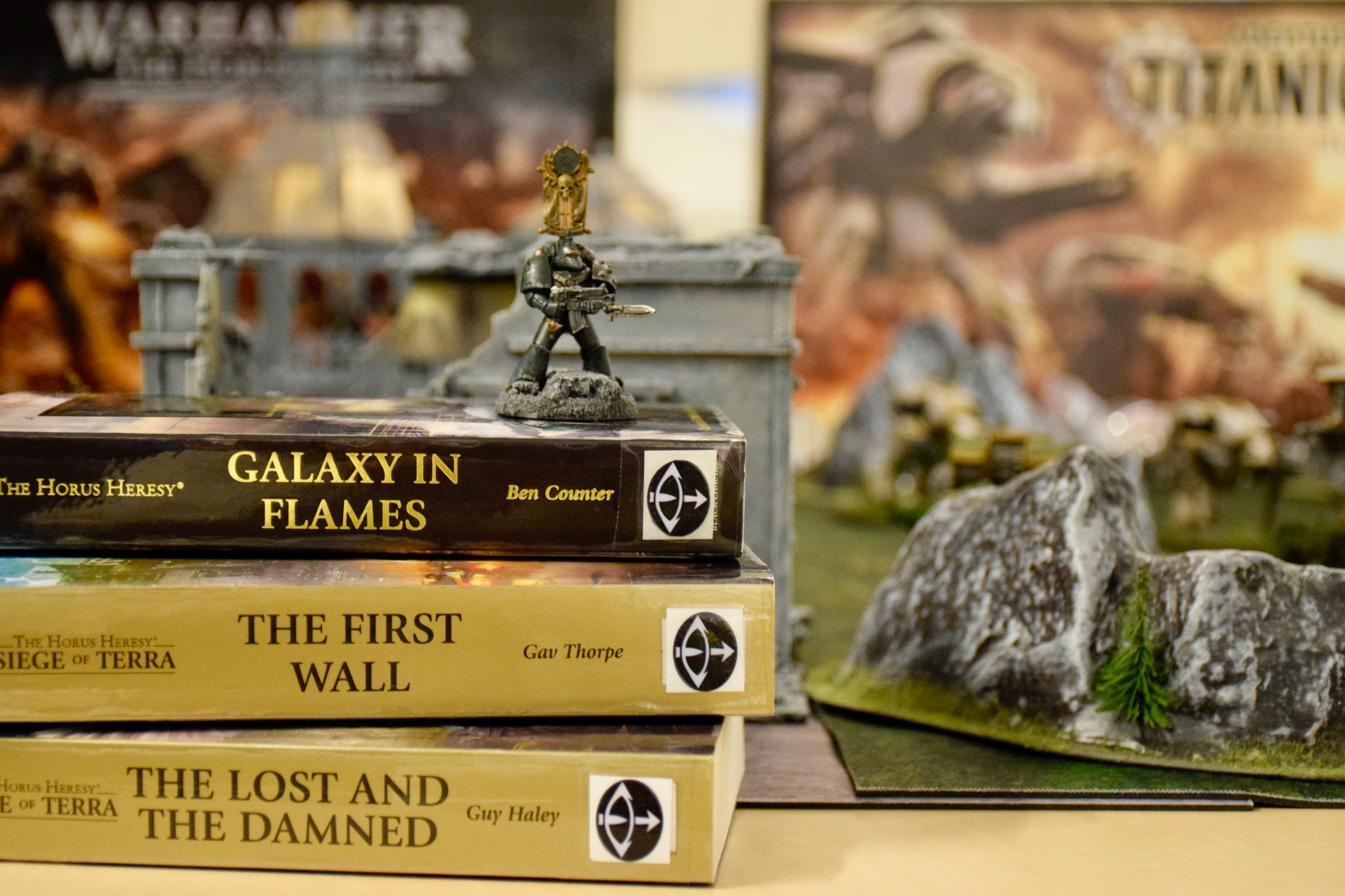 En spelfigur står på en hög med böcker. Böckerna har engelska titlar. Bredvid finns en bergformation som tillhör spelet.