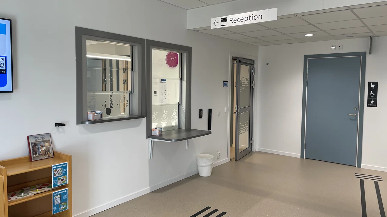 Ett fönster i den vita väggen ut receptionens lucka. I taket hänger en skylt där det står "reception".