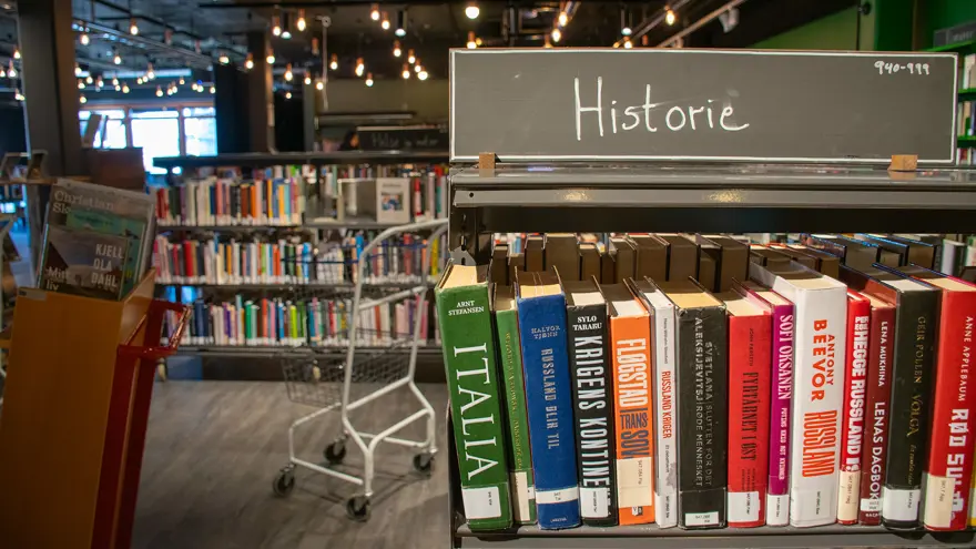 En bokhylla fylld med böcker med skylten "Historie". Bredvid hyllan står en mobil bokvagn och en vagn med två metallkorgar som är tom.