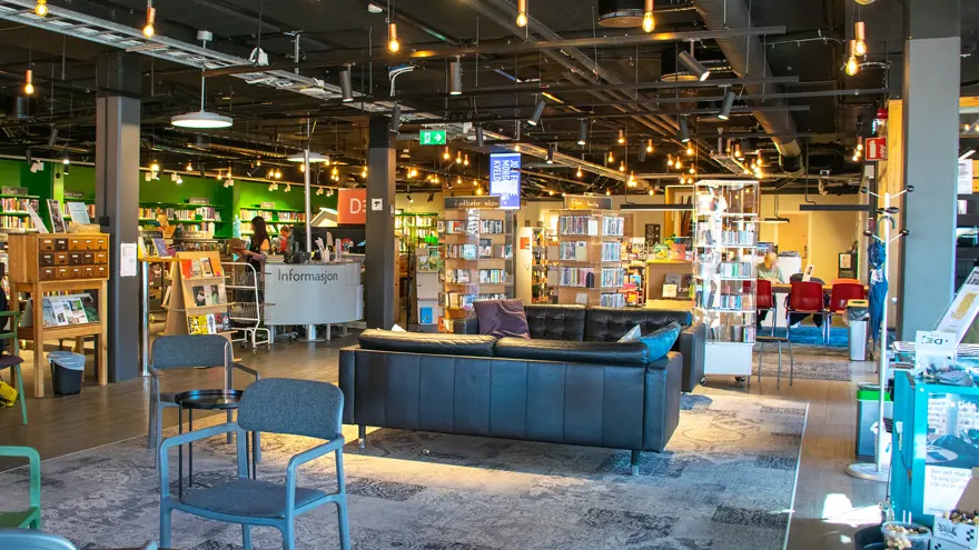 En soffgrupp i svart skinn står i biblioteksrummet. En stor matta ligger på golvet och i taket hänger små spotlights. Informationsdesken finns till vänster om soffgruppen och entrén till biblioteket finns till höger om den.