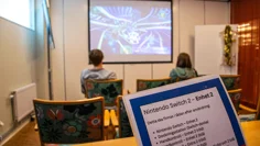 I ett mindre rum sitter en tjej och en kille i tonåren och spelar. De har blickarna på filmduken på väggen där spelet pågår. I förgrunden finns en lapp med instruktioner om Nintendo Switch.