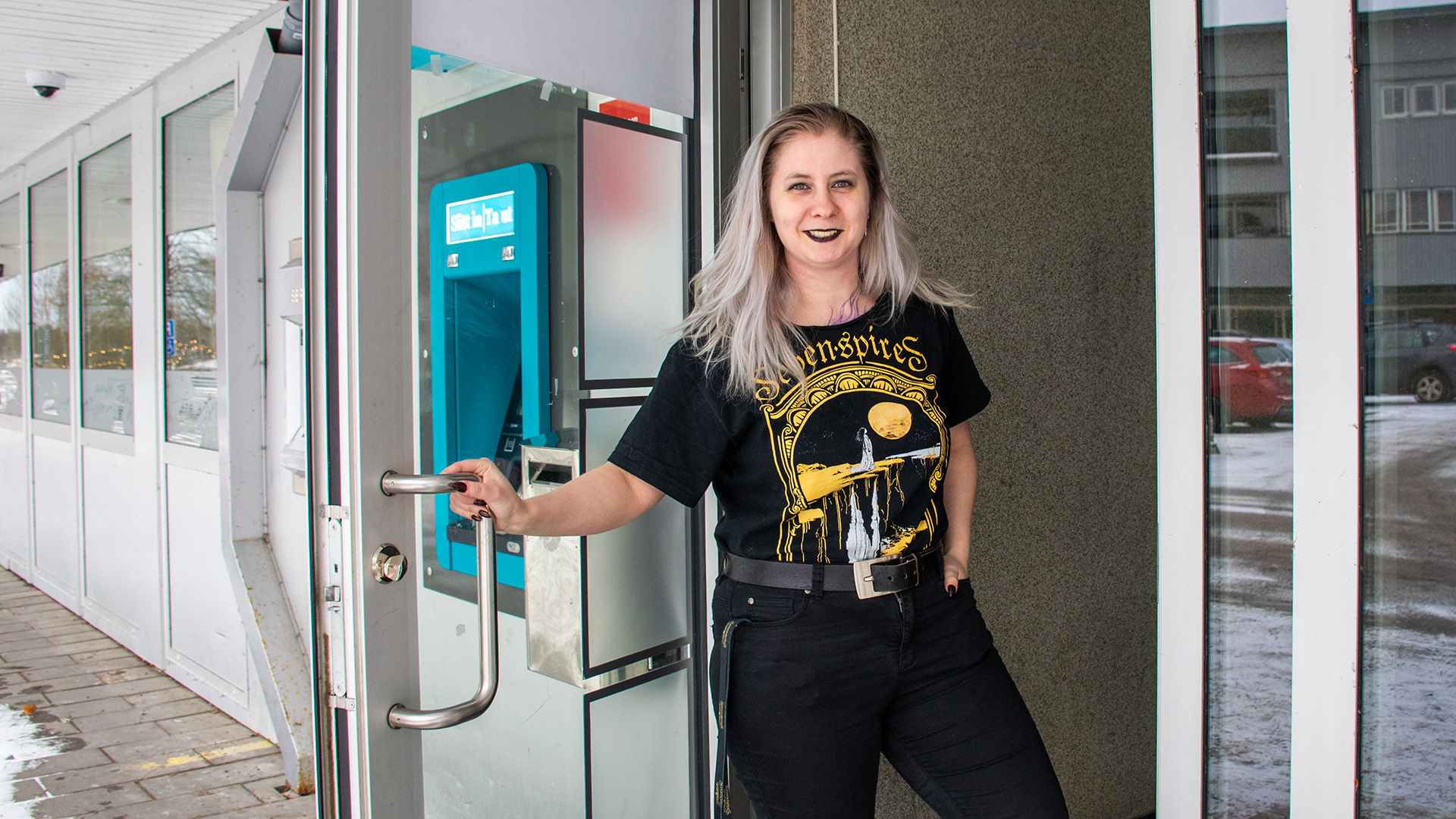Jenny Andersson är i 30-35-årsåldern, hon är klädd i svarta jeans och en svart t-shirt med gult tryck. Hon har långt ljusblont hår och svart läppstift. Hon står i en dörröppning och håller upp dörren medan hon tittar i kameran och ler. Utanför på trottoaren finns snö och en bankomat syns bakom glasdörren hon håller i.