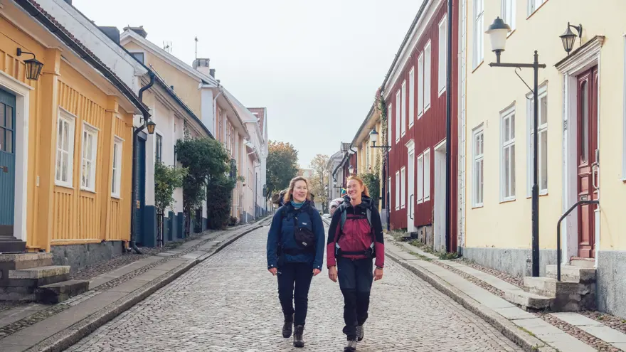 Två kvinnor passerar färgglada trähus på en kullerstensgata.