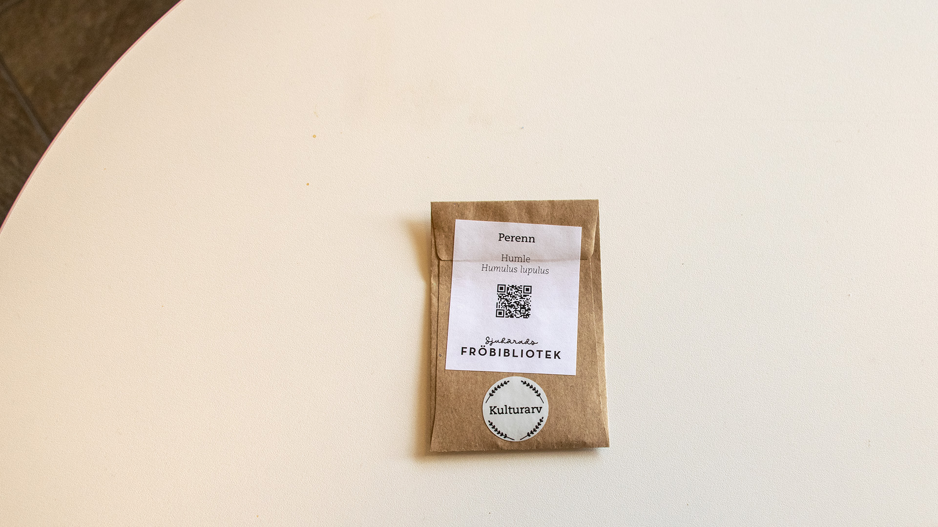 En brun liten fröpåse ligger på ett vitt bord. På den står det bland annat Humle, Perenn och Sjuhärads fröbibliotek. En QR-kod finns också på fröpåsen.