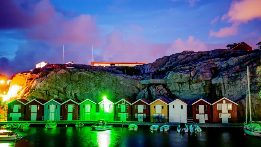 Tolv sjöbodar på rad framför en brygga i skymningsljus.