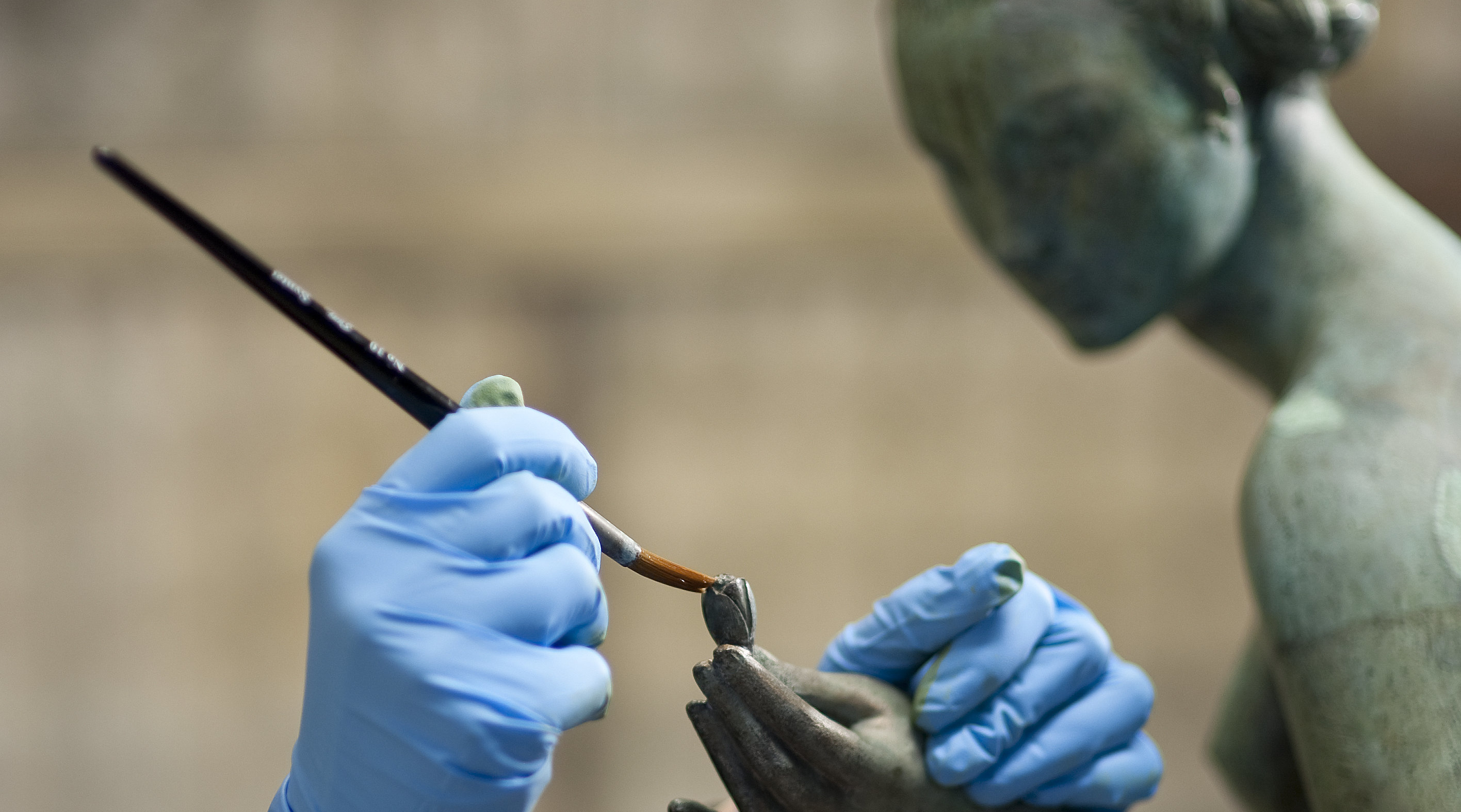 Blåa plasthandskar med en pensel i handen. Arbetar på en bronsstaty, föreställande en naken kvinna. Vi ser huvud, hand och skuldra.