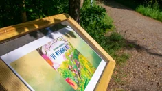 En låg skylt med träram vid sidan av en gångväg i skogen. Innanför glaset finns framsidan till boken Vi rymmer.