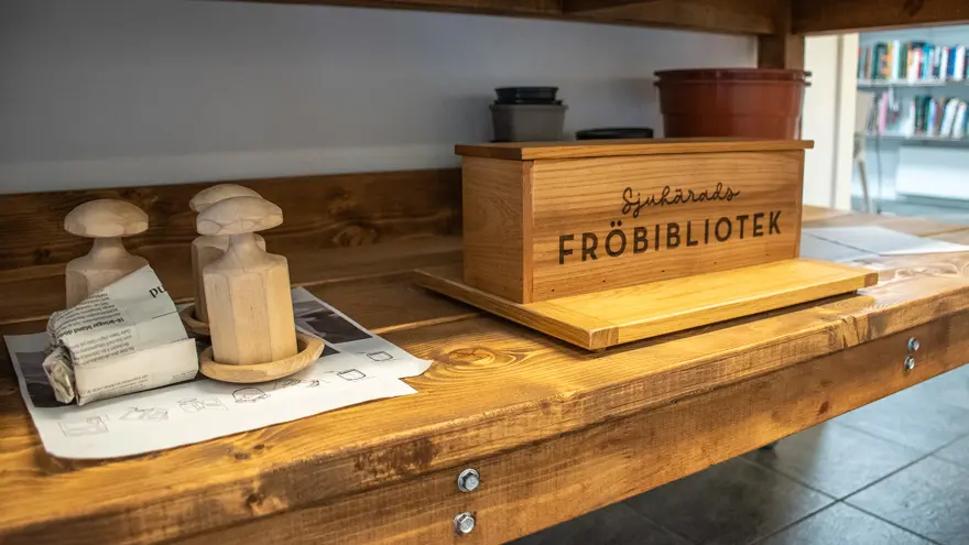 En hylla i trä, på den finns ett träverktyg som man kan göra odlingsurnor i tidningspapper med. En stor trälåda med texten Sjuhärads fröbibliotek och krukor i plast står också på hyllan.