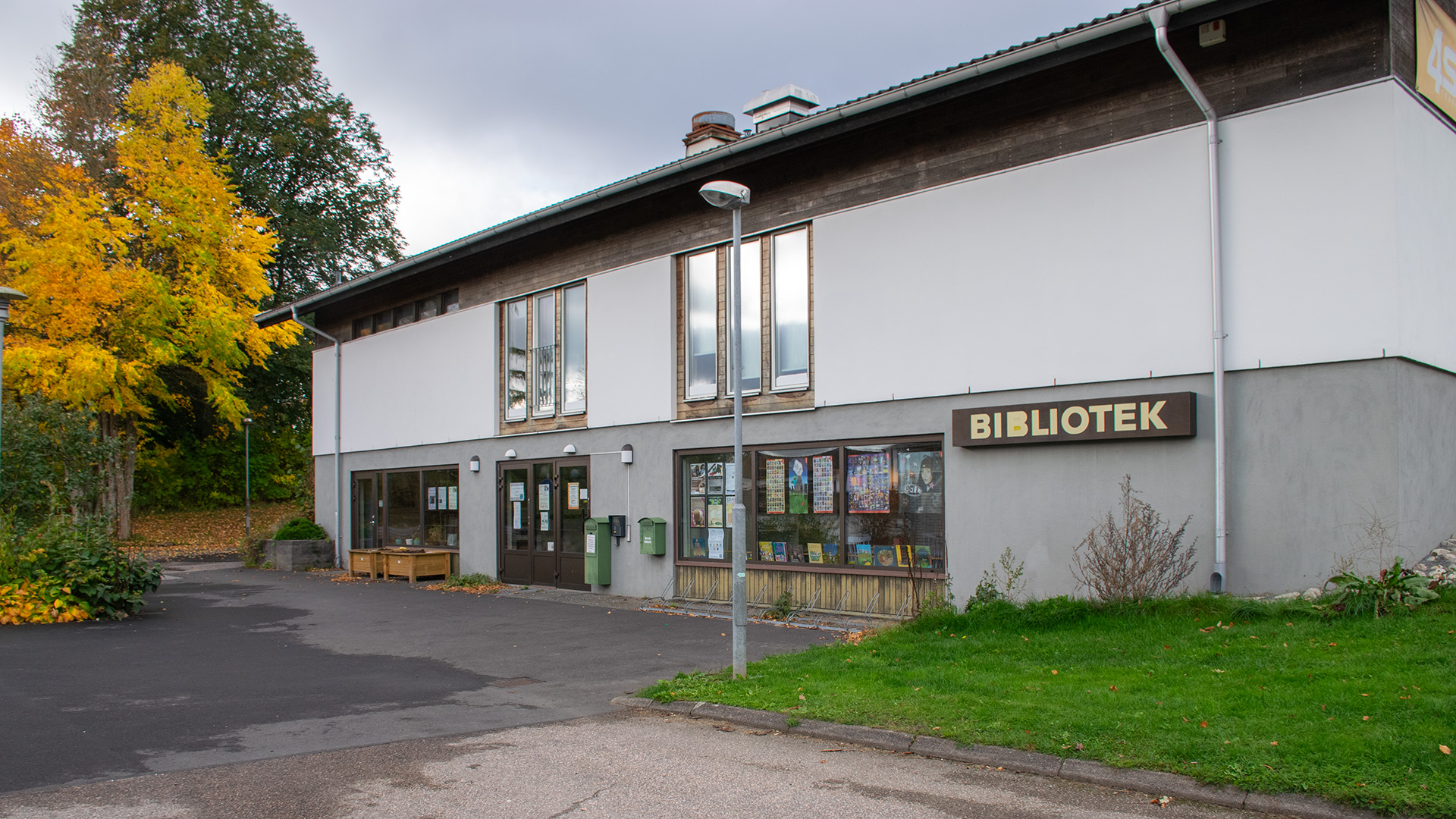Horreds bibliotek. En tvåvåningsbyggnad från gissningsvis 70-talet. Mörkt trä och ljus rappad yta med bruna detaljer runt dörrar och fönster. Utanför biblioteket står två odlingslådor, ett stort träd med gula löv fångar blicken.