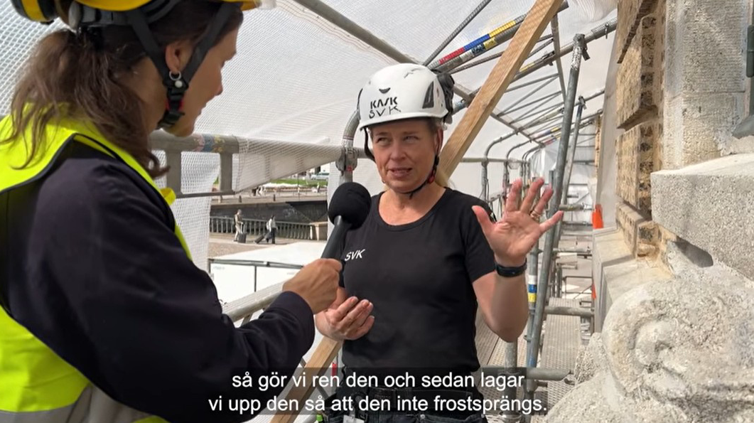 En kvinna i gul reflexväst och svart tröja närmast kameran. Hon håller i en mikrofon. Kvinnan hon pratar med har en vit hjälm på huvudet och svart t-shirt. De står intill en beige fasad under inplastade byggställningar