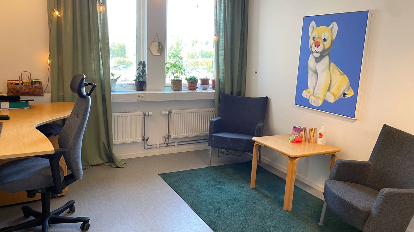 Mottagningsrum med två fåtöljer, skrivbord och en tavla på väggen.