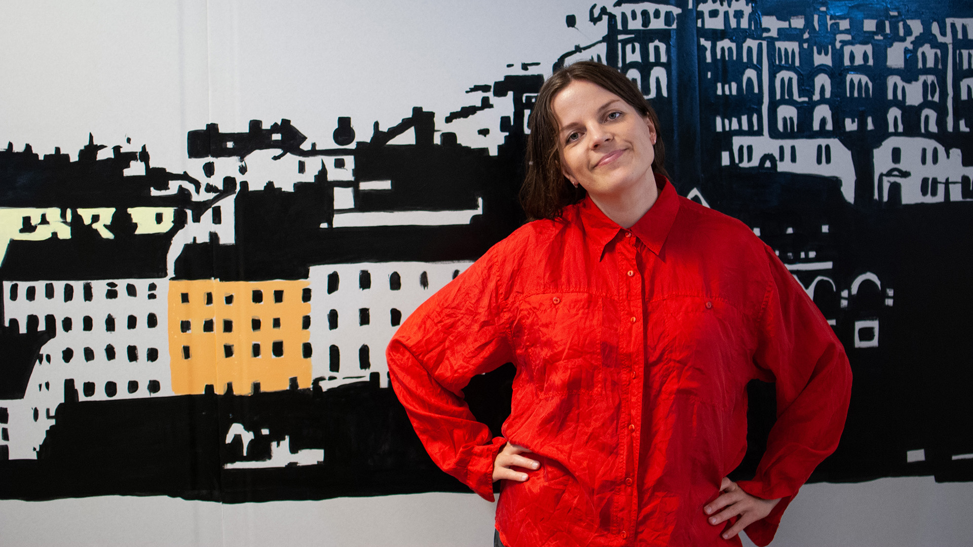 Milla Leskinen är i 35–40-årsåldern. Hon har mörkt hår som är samlat i en tofs och är klädd i en klarröd skjorta. Hon står mot en vägg med ett målat stadsmotiv, hon har händerna i midjan, huvudet lite på sned och ler med stängd mun.