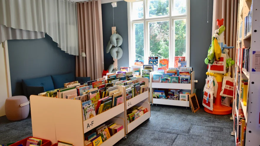 Barnavdelningen på biblioteket. Låg bokhyllor i barnhöjd står i mitten av rummet och vid fönstret. En blå soffa och en sittpuff står i ett hörn.