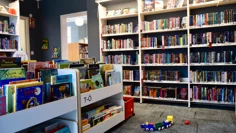 Barnavdelningen på biblioteket. På golvet finns en heltäckningsmatta, väggarna är målade i en dov gråblå färg. Bokhyllor utmed väggarna och boklådor i mitten av rummet och på golvet står ett färgglatt leksakståg i plast.