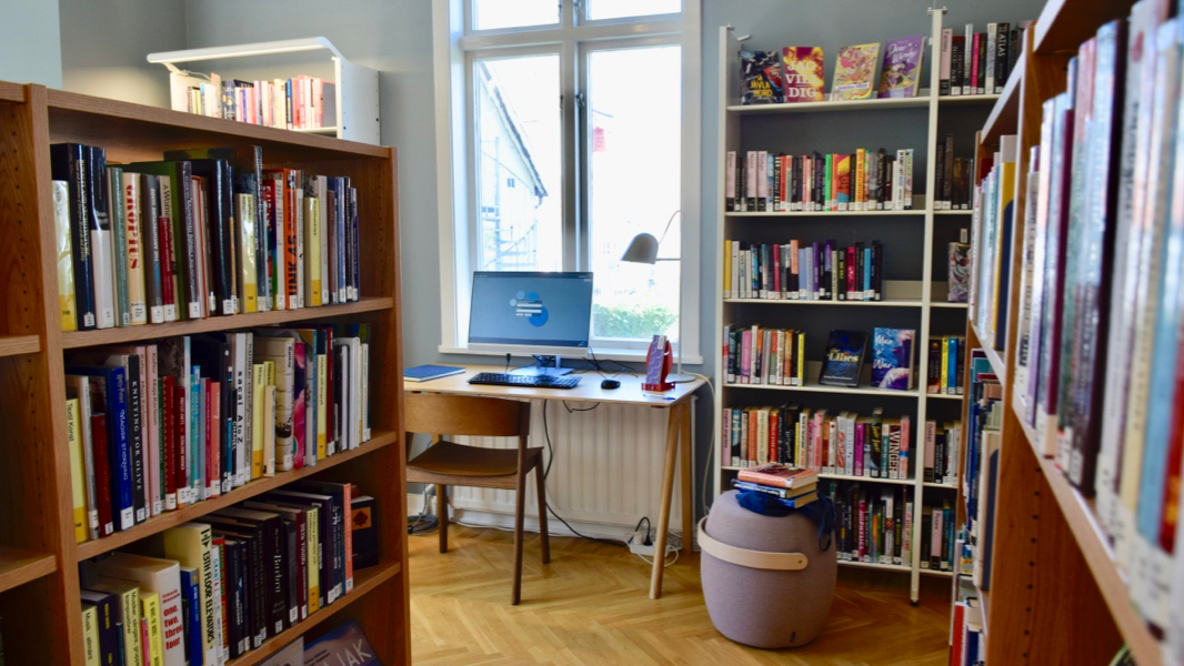 Fyllda bokhyllor inne på vuxenavdelningen. Vid ett fönster står ett litet skrivbord med en datorskärm och en stol. 
