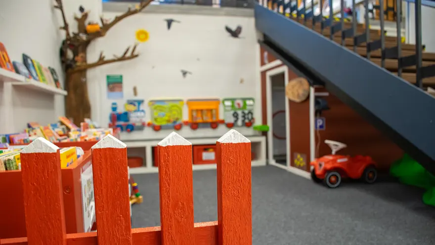 Ett rött staket i förgrunden. Hela Sagohult finns bakom det, boktråg, snickarboa, en bobbycar, en bänk är några saker som finns i den färgglada hörnan.