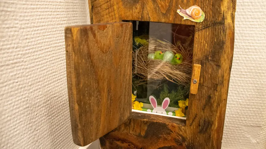 I ett stiliserat träd finns en lucka. Luckan är öppen och innanför finns ett litet fack med ett fågelbo med små gröna småfågelungar i.