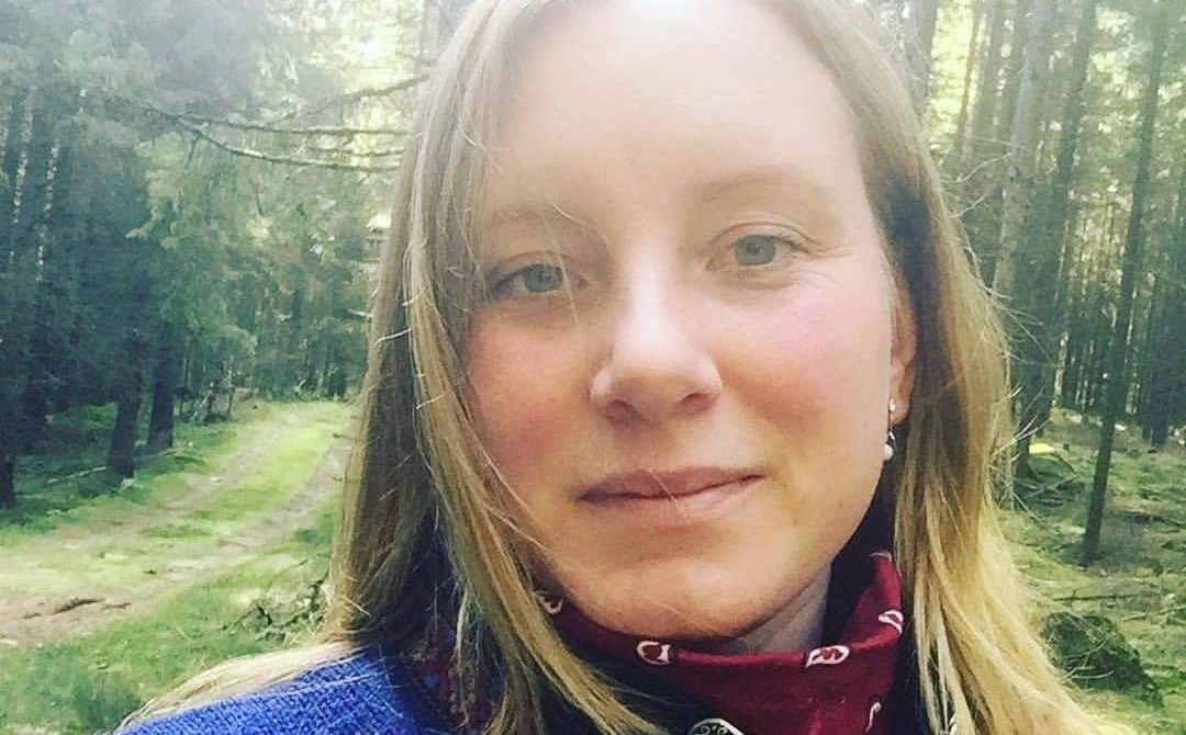 Personen tittar in i kameran och ler. Personen står i skogen i dagsljuset.