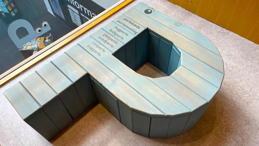 En stor träskulptur i formen som ett stort P. Den ligger plant mot golvet och på den står ord skrivna som innehåller bokstaven P.