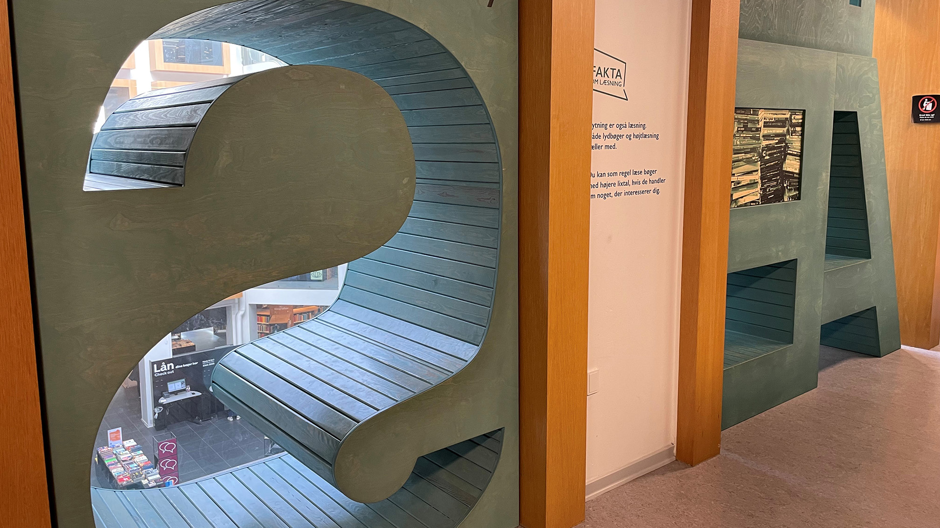 En sittplats i biblioteket, den är formad som bokstaven S och har utsikt ner mot våningen nedanför. Lite längre bort finns fler sittplatser och hyllor utformade som bokstäver. 