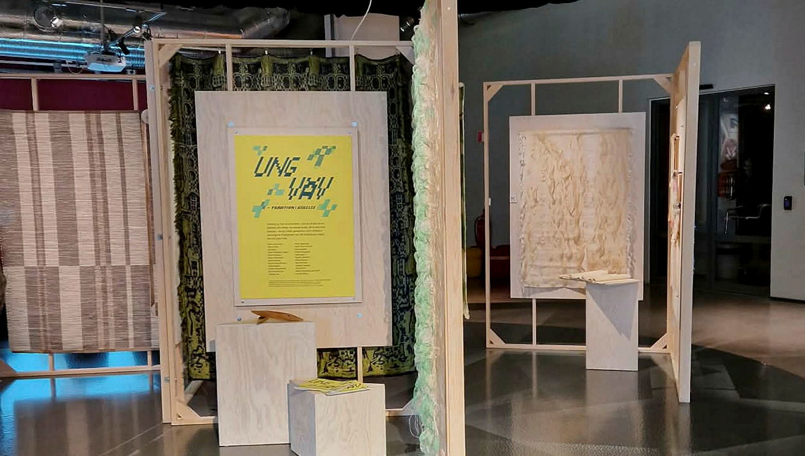 Utställningen UNG VÄV på textilmuseet i Borås. Från vänster: en stor matta vävd med stora rutor i grått och naturvitt. I mitten syns två stora träramar i vinkel. på den som är rakt fram sitter en gul affisch med texten UNG VÄV i grönt och bakom den syns en stor grön väv mes svart mönster. Framför affischen finns två podier med kataloger på. I ramen som kommer rakt mot mitten anas en blöjande väv i ljusgrönt och vitt. Till höger finns ytterligare en vinkel med träramar. På den ena syns en naturvit väv med lång lugg och framför den ett podium med en bok av tyg.